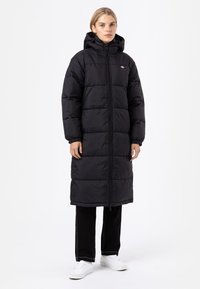 Black puffer coat with a hood, quilted design, full zipper closure, knee-length, and loosely fitted sleeves. Worn with black pants and white shoes.