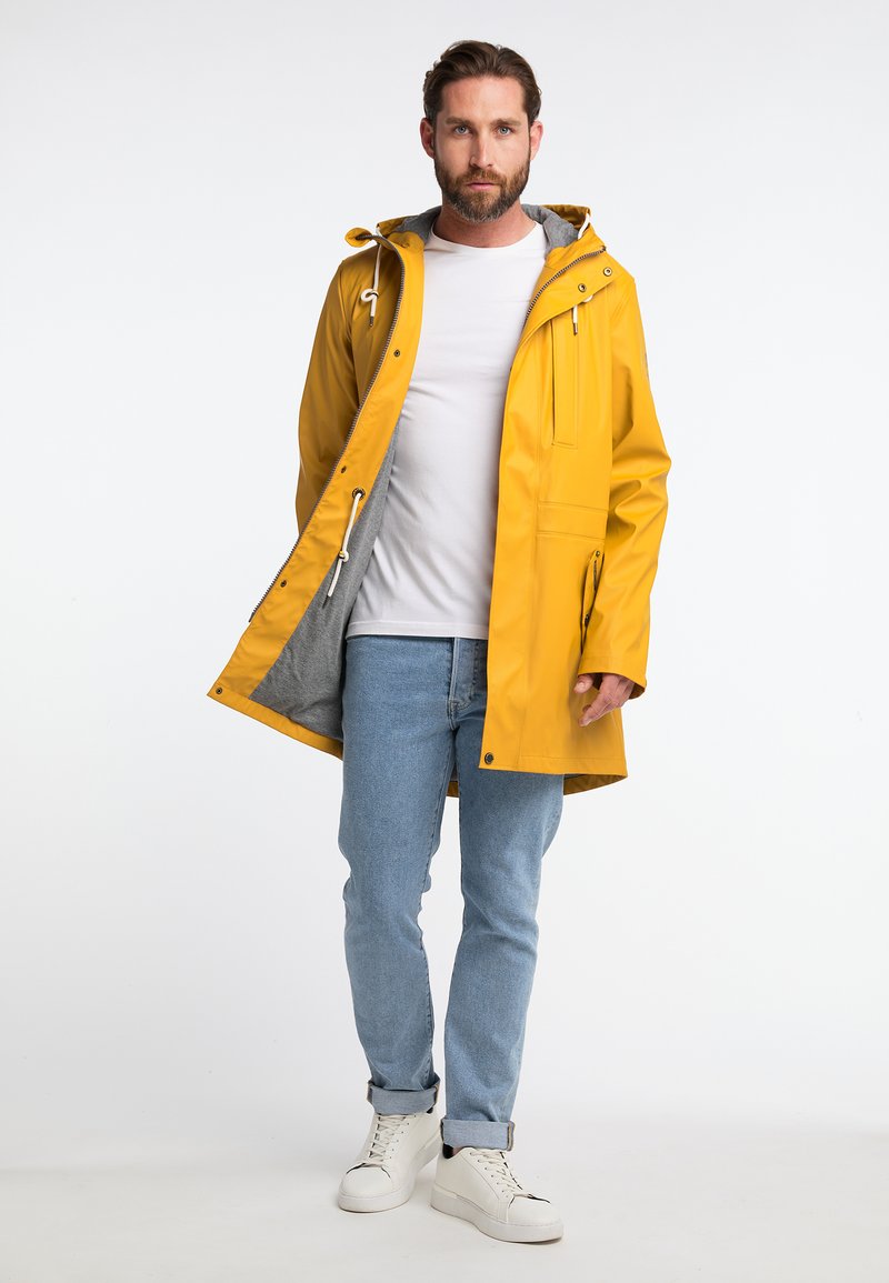 Gelbe wasserdichte Regenjacke mit grauem Fleece-Futter, verstellbarem Kordelzug im Kapuzenbereich und Fronttaschen. Kombiniert mit blauen Jeans und weißen Turnschuhen.