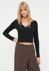 Čierny dlhý rukáv cropped top s véčkovým výstrihom a tromi gombíkmi na výstrihu, skombinovaný s hnedými nohavicami s vysokým pásom. Hladká textúra.