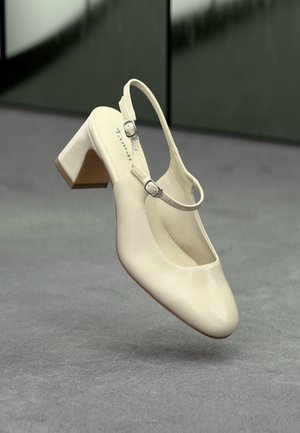 Cream-colored leather slingback shoe with low block heel and two adjustable silver buckles on gray floor background.
