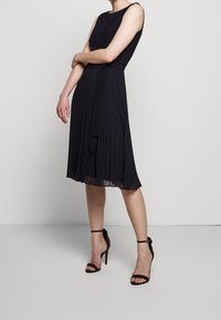 Žena v černých bezramínkových plisovaných midi šatech s páskem v pase, doplněn�á černými sandály na vysokém podpatku s páskem kolem kotníku.