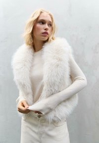 Weißer Kunstfell-Westen über einem beigen Pullover, der lange Ärmel und eine strukturierte Oberfläche hat. Das Modell hält den Pullover am Saum.