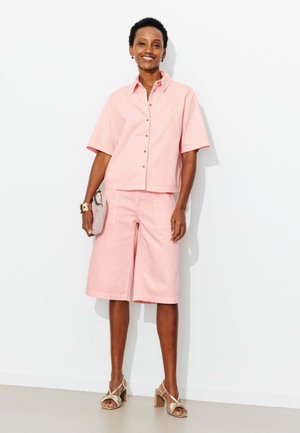 Femme souriante portant une chemise rose clair à manches courtes avec boutons et un short assorti jusqu'aux genoux, tenant une petite pochette beige.