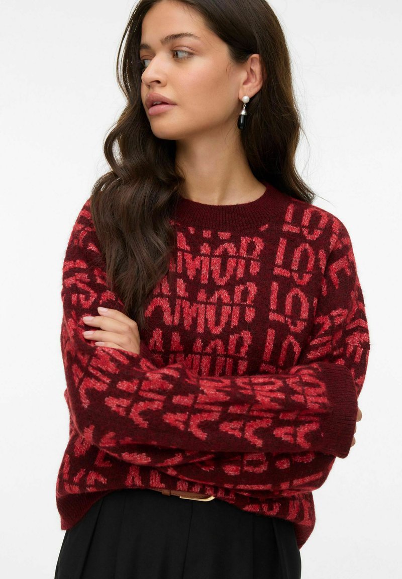 Pull en tricot rouge avec un motif de texte répété "AMOUR" dans des teintes variées. Coupe ample avec des poignets côtelés et un col rond.