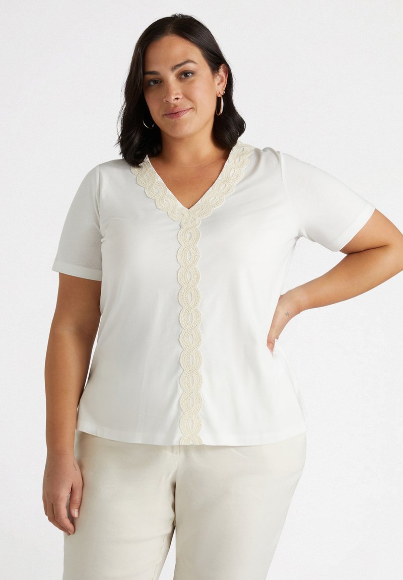 Top blanco de manga corta con escote en V y ribete de encaje crema a lo largo del cuello. Tejido suave y corte relajado, combinado con pantalones de color claro.