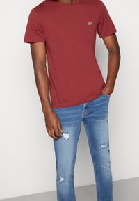 Maroon cotton t-shirt with short sleeves and round neck, featuring a small green logo. Paired with light blue distressed jeans.