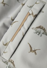 Una tela de color gris claro presenta un patrón de dinosaurios, incluyendo un estegosaurio y un pterosaurio, con una textura sutil y una costura de acento en color arena.