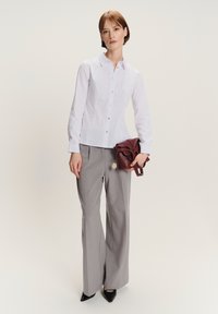 Donna che indossa una camicia bianca con i bottoni e pantaloni ampi grigi, tiene in mano una borsa piegata color bordeaux, in piedi su uno sfondo neutro.