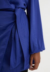 Blaues Wickeloberteil aus leichtem Stoff, mit gebundenem Taillengürtel, langen puffärmel und überlappendem Design mit weicher Textur.
