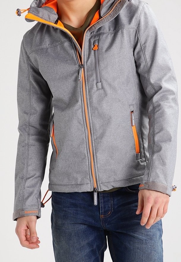 Grijze jas met oranje accenten, ritszakken en verstelbare capuchon. Heeft een getextureerd materiaal en opvallende stikdetails.