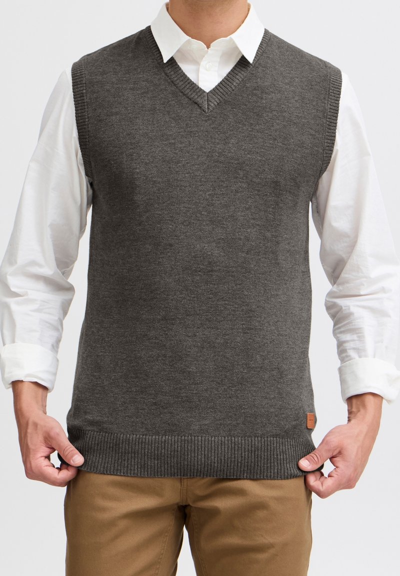 Szary sweter bez rękawów z dzianiny z dekoltem w kształcie litery V, ściągaczem u dołu i bocznymi akcentami. Noszony na białej koszuli zapinanej na guziki, o gładkiej fakturze.