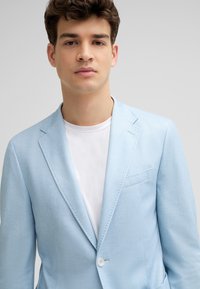 Helles blauer Blazer aus strukturiertem Stoff mit fransigem Revers, einem weißen Knopf und einer Brusttasche. Über einem weißen T-Shirt getragen.