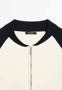 Indumento bianco con collo e maniche a coste nere, cerniera centrale argentata e etichetta "Massimo Dutti Made in China" all'interno del collo.