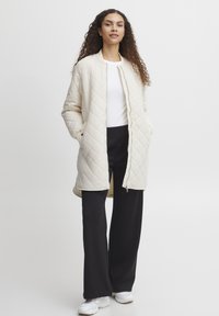 Ljus beige kviltad jacka med långa ärmar, framdragkedja och sidofickor, tillsammans med en vit topp och svarta byxor. Modell som står.