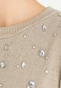 Beige gebreide trui met verschillende maten heldere strass steentjes verspreid over de stof, ronde halslijn en een gestructureerd breipatroon.