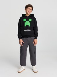 Czarna bluza z kapturem z zielonym wzorem twarzy Minecraft Creeper, posiadająca przednią kieszeń i mankiety; w komplecie szare spodnie typu cargo i białe tenisówki.