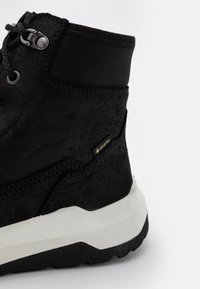 Černá voděodolná bota s texturovaným semišovým povrchem, kulatými tkaničkami a zesíleným kotníkem. Obsahuje bílou mezipodešev a embosované detaily značky.