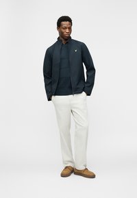 Νεαρός άνδρας όρθιος με τα χέρια στις τσέπες, φοράει μπλε σκούρο μπουφάν, μπλε σκούρο πουκάμισο, λευκό παντελόνι και καφέ παπούτσια σουέτ, μπροστά σε λιτό φόντο.