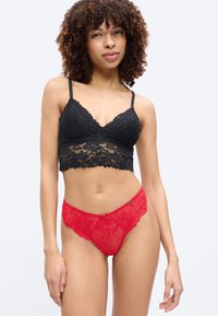 Γυναίκα με σγουρά μαλλιά που φοράει μαύρο δαντελένιο σουτιέν και κόκκινο δαντελένιο εσώρουχο, στέκεται μπροστά σε απλό λευκό φόντο.