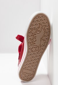 Semelle d'une basket rouge montrant une bande de roulement en caoutchouc marron avec un motif de chevaux et d'étoiles, une semelle intermédiaire blanche, et le logo "MUSTANG".