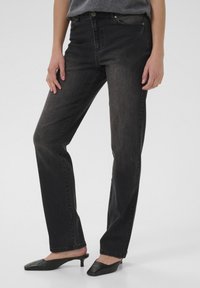 Osoba v zažloutlých černých rovně padnoucích džínách a černých střevících s malým podpatkem, stojící na jednobarevném světlém pozadí.