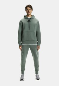 Zöld kapucnis pulóver elülső zsebbel, összeillő joggingnadrággal párosítva. Fekete húzózsinórral és texturált anyaggal ellátva.