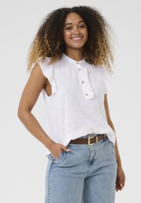 Weißes Leinenoberteil mit rüschigen Ärmeln und einer Knopfleiste vorne. Kombiniert mit blauen High-Waist-Jeans und einem braunen Gürtel. Die Textur wirkt leicht.