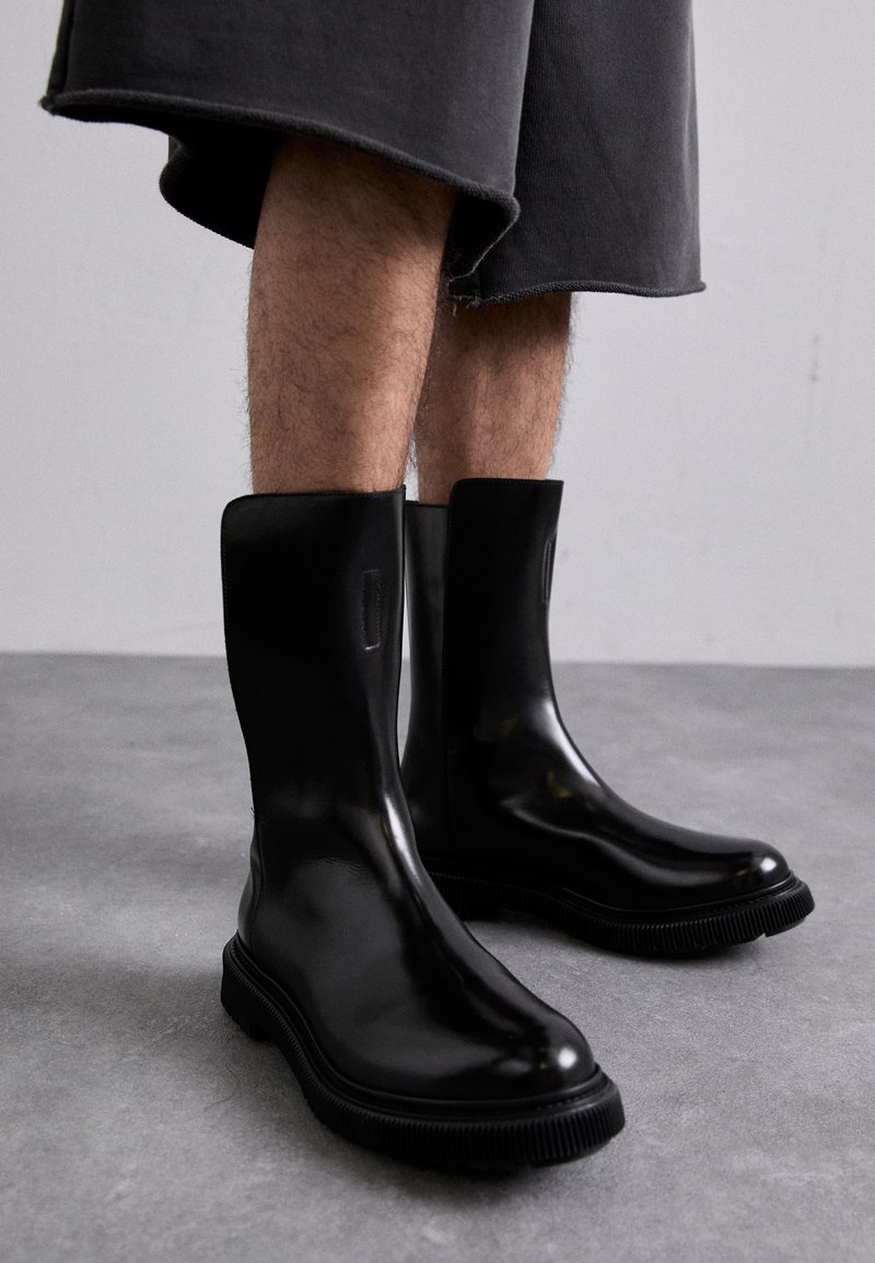 Μαύρες καουτσούκ μποτίνες μέχρι τον αστράγαλο με λεία επιφάνεια, υφαντή σόλα με ραβδώσεις και ελάχιστες λεπτομέρειες, σε συνδυασμό με γκρι σορτς.