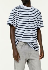 Person, der ein weißes und navyblau gestreiftes Kurzarmshirt trägt, das teilweise in eine hellgraue Jeans gesteckt ist, und vor einem schlichten Hintergrund steht.