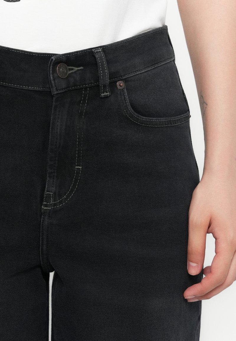 Jean en denim noir taille haute avec des accents décolorés, présentant une fermeture à bouton, un design à cinq poches, et des détails de couture contrastés.
