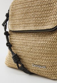 Vevd stråcrossbody-veske i beige med en teksturert overflate. Har en svart glidelås og justerbar svart stropp. Merkevaren er synlig.