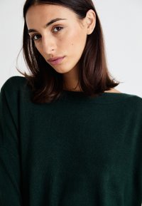 Pulover verde tricotat, cu o croială lejeră și decolteu larg. Textură moale cu un luciu subtil, având umeri căzuți pentru un stil relaxat.