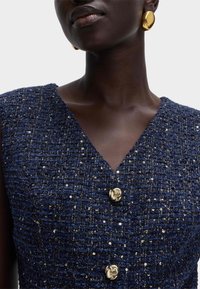 Marineblauwe jurk met textuur en pailletten en een V-hals, voorzien van twee gouden ronde knopen als accenten. Glad materiaal met fijne details.