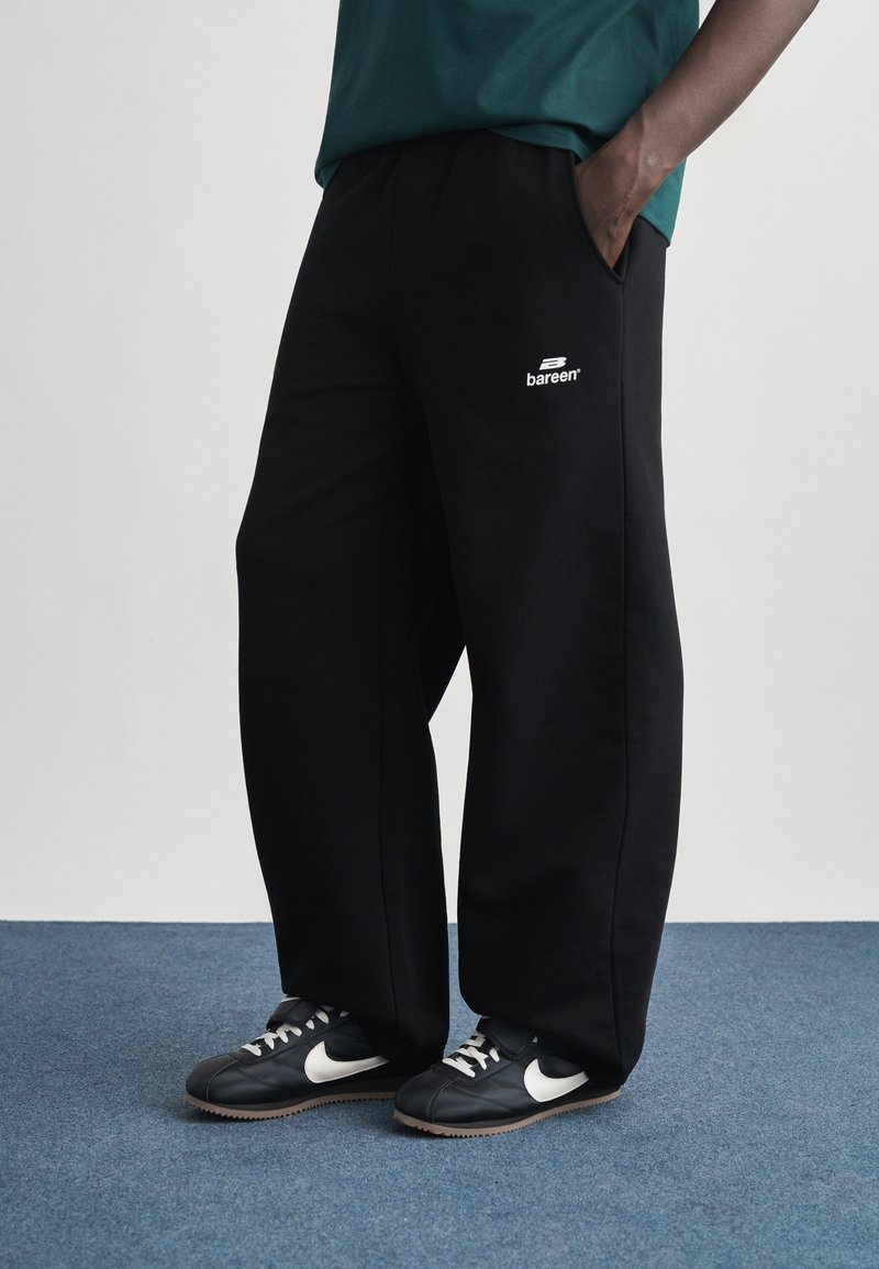 Άτομο που φοράει μαύρο φαρδύ παντελόνι φόρμας με λευκό λογότυπο "bareen" και μαύρα παπούτσια Nike με λευκές γραμμές σε μπλε χαλί.