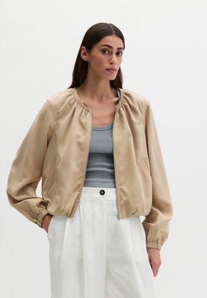 Frau mit langen dunklen Haaren trägt eine beige Jacke mit Reißverschluss über einem grauen Tanktop und weißen hoch taillierten Hosen, steht vor einem schlichten Hintergrund.