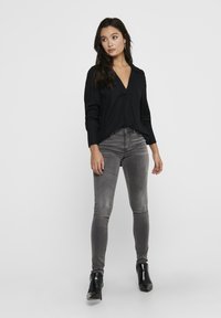 Černé dlouhé rukávy tričko s výstřihem do V a volným střihem, kombinované se šedými skinny džíny. Modelka nosí černé kotníkové boty. Jednoduché pozadí.