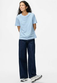 Camisa de manga corta azul claro con lunares negros, combinada con pantalones de pierna ancha de denim oscuro y zapatillas deportivas plateadas.