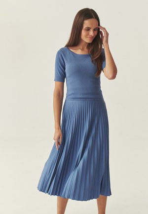 Woman in blue short-sleeve top and matching pleated midi skirt, gently touching her hair, standing against a plain light background.
