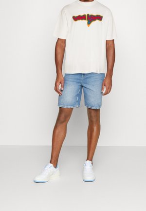 Man som bär en vit t-shirt med färgglad "VON LASH" text, ljusblå denimshorts och vita sneakers med ljusblå sulor.