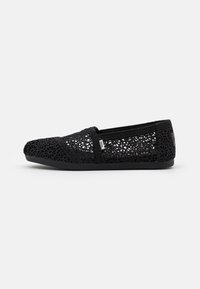 Chaussure noire en crochet à enfiler avec un motif en dentelle texturé, un bout arrondi et une semelle en caoutchouc noir flexible. Comprend des accents de couture et une étiquette avec le logo.
