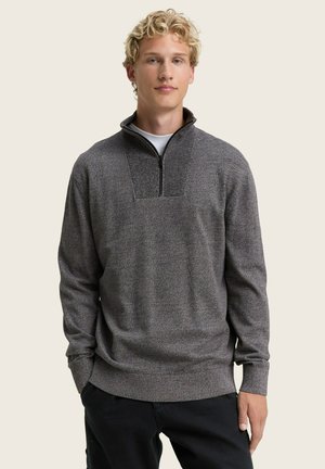 Grauer Reißverschluss-Pullover mit strukturiertem Design, hohem Kragen und geripptem Saum sowie Ärmeln. Über einem weißen T-Shirt mit lockerer Passform getragen.