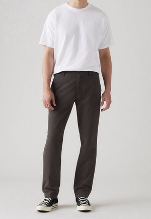Mann trägt ein schlichtes weißes T-Shirt mit kurzen Ärmeln, dunkelbraune gerade geschnittene Hose und schwarze Sneakers mit weißen Schnürsenkeln und Sohlen.