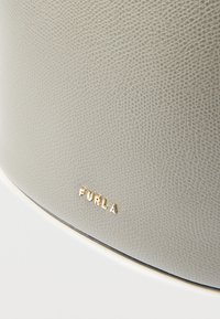 Geantă de mână din piele texturată gri, cu logo auriu "FURLA". Prezintă un design rotund și un stil minimalist, accentuând calitatea materialului.