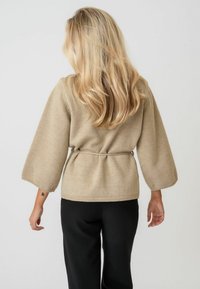 Beige gestrickter Pullover mit langen, weiten Ärmeln und einem Bindeband in der Taille. Getragen mit schwarzen Hosen, die eine schlichte, strukturierte Rückansicht zeigen.