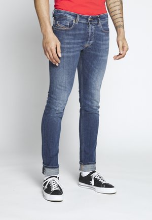 Homme portant un jean slim bleu retroussé aux ourlets, des baskets noires et blanches, des chaussettes blanches et une chemise rouge avec un bras tatoué visible.