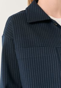 Marineblauwe button-up shirt met een kraag, verticale witte streepjes en een borstzak, gemaakt van textuurstof.