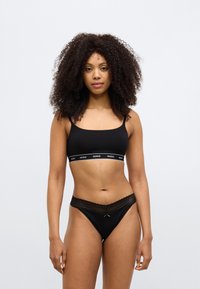 Crni bralette s širokom elastičnom trakom na kojoj je bijeli logotip "HUGO". Odgovarajuće crne tange s čipkastim obrubom i mašnom na pojasu.