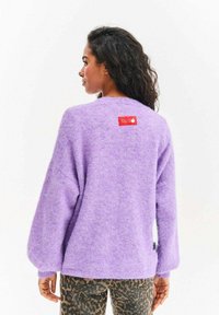 Kobieta z długimi kręconymi włosami, ubrana w puszysty lawendowy sweter z czerwoną metką na plecach i spodnie w panterkę, zwrócona w lewo.