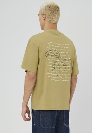T-shirt en coton olive avec une coupe décontractée ; le dos présente un texte blanc et un dessin au trait d'un champignon. Manches courtes, col rond.