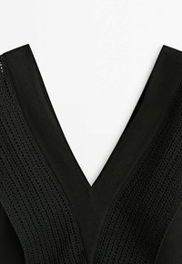 Vêtement noir en V avec une large bordure en tricot texturé le long du col sur fond blanc.